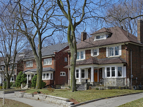 Row of large old detached brick houses with mature trees in the front yard