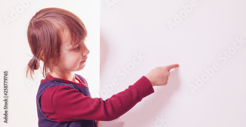 Portrait of little cute child girl behind white empty banner pointing with her finger towards copy space area.