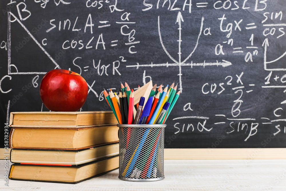 School desk in classroom, with books on background of chalk board with written formulas. Soncept Teacher's Day. School desk in classroom, with books on background of chalk board with written formulas. Soncept Teacher's Day.
