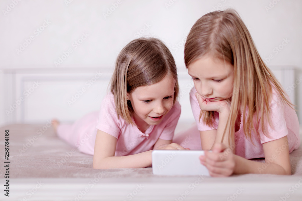 Two little girls (sisters) in pink pajamas on the bed watching a smartphone and reading an e-book. Two little girls (sisters) in pink pajamas on the bed watching a smartphone and reading an e-book.