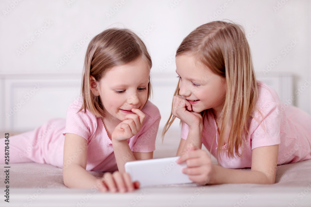 Two little girls (sisters) in pink pajamas on the bed watching a smartphone and reading an e-book. Two little girls (sisters) in pink pajamas on the bed watching a smartphone and reading an e-book.