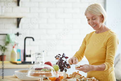 Positive attractive mature woman with blond hair standing at counter and buttering slice of brad while preparing bruschetta in kitchen