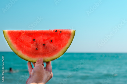 Watermelon, juicy, red slice in the hands of a woman against the background of the ocean, in the rays of sunlight. Diet of tropical fruits.