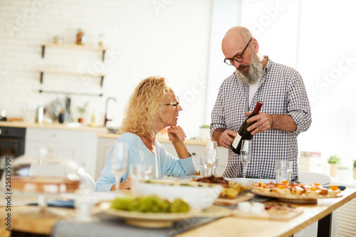 Serious handsome senior bearded man in glasses pointing at ladle of bottle and talking about wine to lady while they having dinner at home