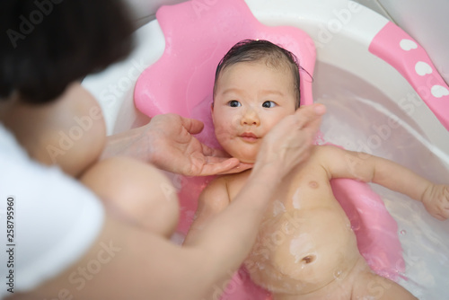 Little young cute Asian baby has been taking a bath in bathtub and washing by her mother. She is looking and her mother when mother touch her cheek. Baby health care concept.