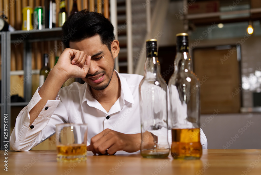 A man sitting at the pub is crying due to his sadness and want to forget it by drinking alcohol. He drank a lot of whiskey, he finished one bottle and continue on second bottle. Alcoholic concept.