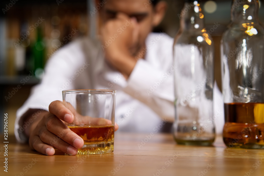 Drunk man sitting at the bar in pub and holding whisky glass in right hand and put another hand on his head after he drank a lot of alcohol and feeling headache. Alcoholic concept. Drunk man sitting at the bar in pub and holding whisky glass in right hand and put another hand on his head after he drank a lot of alcohol and feeling headache. Alcoholic concept.