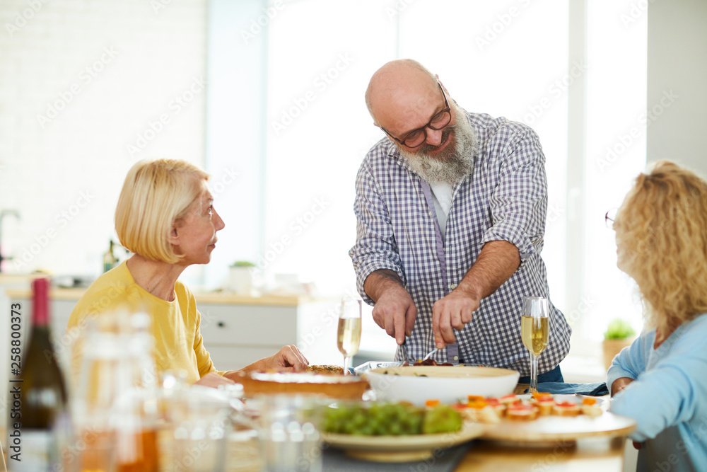 Content handsome mature bearded man in glasses standing at table and cutting meat for guests while talking to charming ladies at home