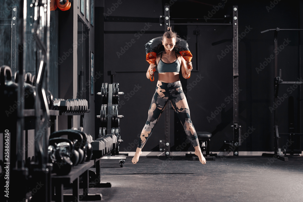 Fototapeta premium Full length of Caucasian female bodybuilder jumping with bulgarian sack on shoulders. Night workout in gym concept.