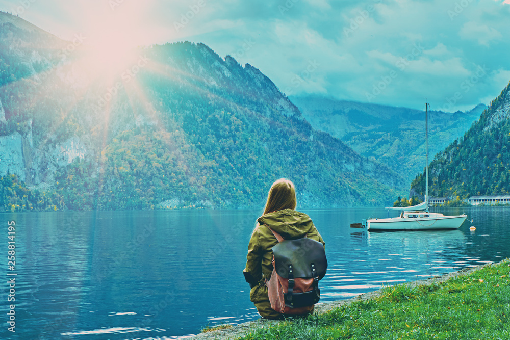 Young tourist girl with backpack looking at the beautiful scenic sunset at Austrian alps lake. Hipster travel vacation in alps mountains concept. Nice yacht boats on calm peaceful alps lake. Toned
