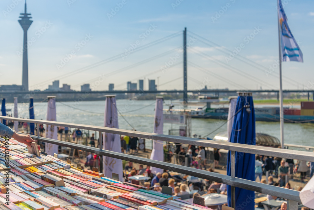 Stack of books of open air book store and blurry view of peoples in outdoor sunny restaurant at promenade along riverside of Rhine River and background of downtown.