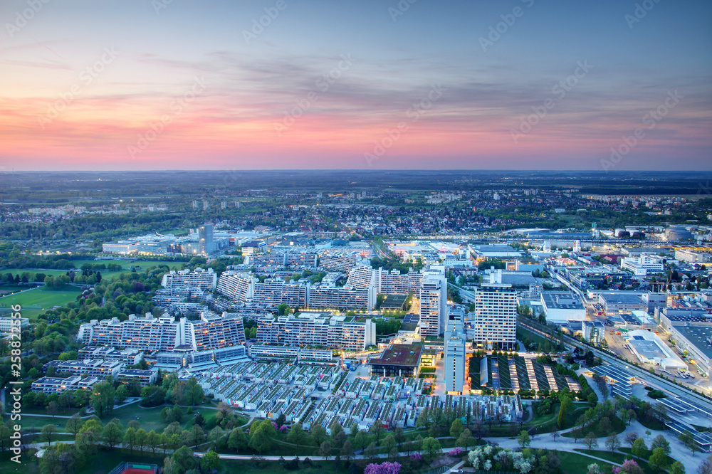 Modern European city outskirts aerial view in blue hour at dusk, concrete high-rise buildings and parks of dense housing complex with industrial commercial area in background, Munchen Germany Europe