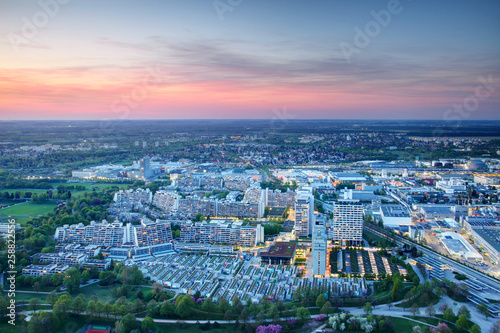 Modern European city outskirts aerial view in blue hour at dusk, concrete high-rise buildings and parks of dense housing complex with industrial commercial area in background, Munchen Germany Europe