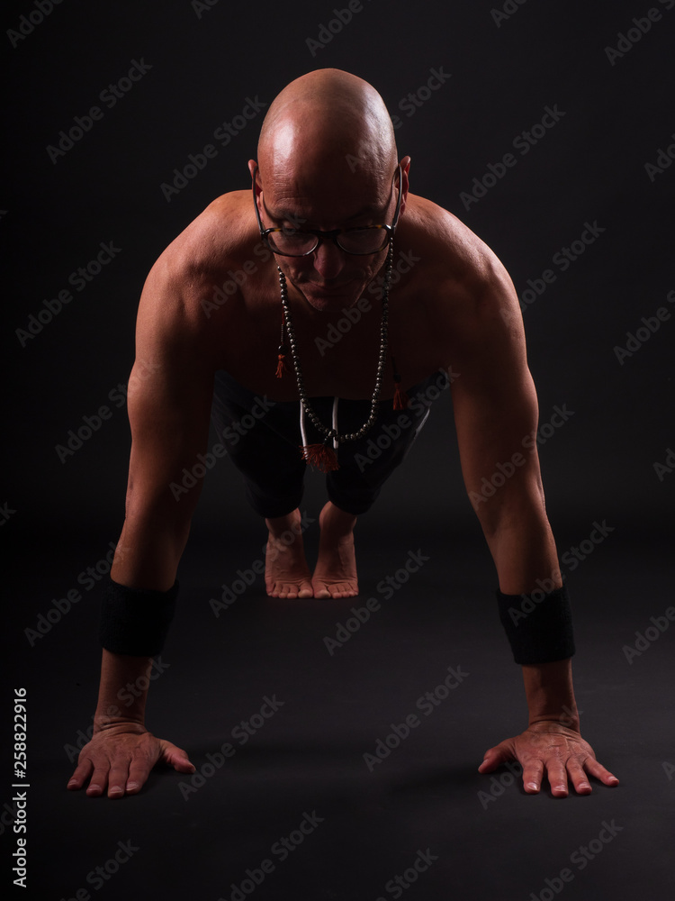 Buddhist monk practicing yoga on black background in long pants and glasses
