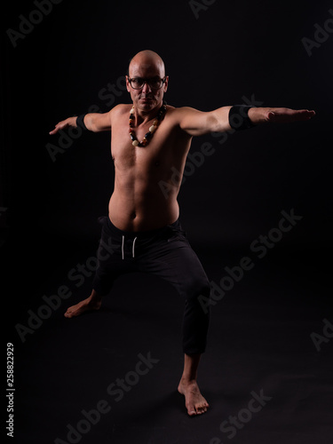 Buddhist monk practicing yoga on black background in long pants and glasses