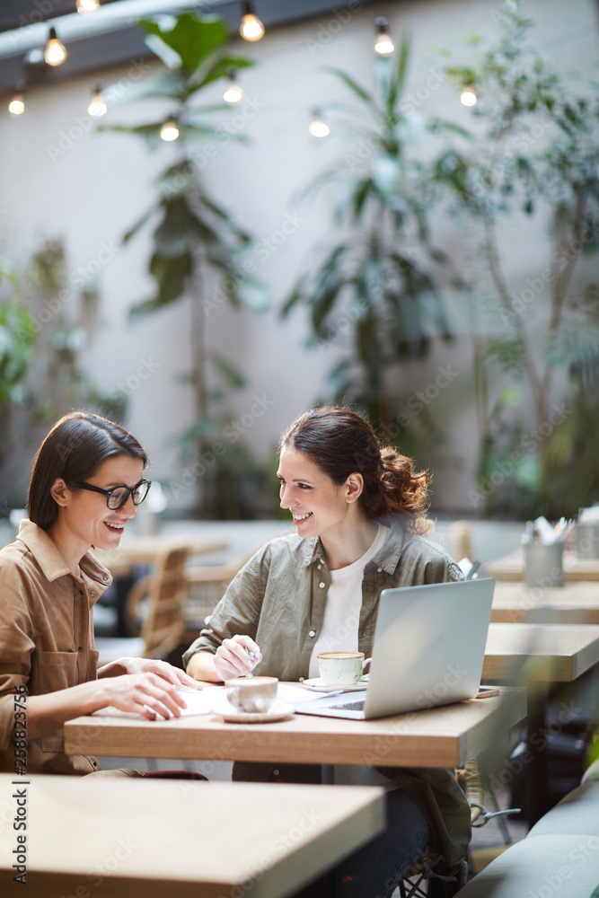 Cheerful excited female entrepreneurs in casual shirts sitting at table in modern coffee shop and elaborating strategy while analyzing papers