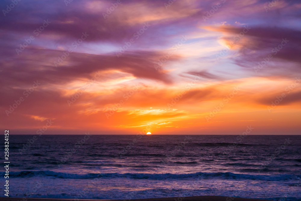 Sun disappears beneath horizon over ocean after spectacular sunset with vivid sky. Wide angle scene at dusk.