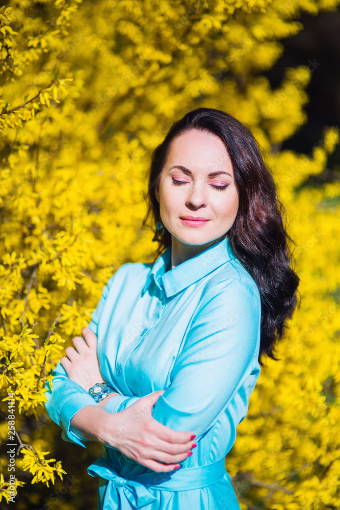 portrait of a brunette girl with closed eyes in a blue dress at a blooming yellow Bush