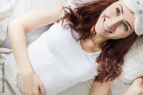 An attractive young brunette girl with brown hair wakes up in her bed in a sleep mask. View from above