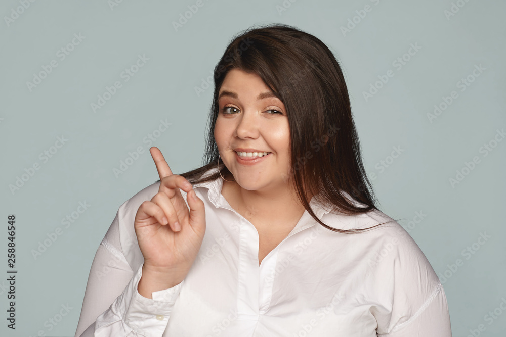 Check this out. Playful beautiful young female with loose black hair and extra pounds winking at camera and smiling, pointing fore finger away at blank wall with copy space for your content Check this out. Playful beautiful young female with loose black hair and extra pounds winking at camera and smiling, pointing fore finger away at blank wall with copy space for your content