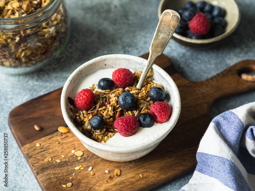 A white bowl of homemade granola, yogurt, fresh berries on wood board. Vegetarian food, healthy breakfast