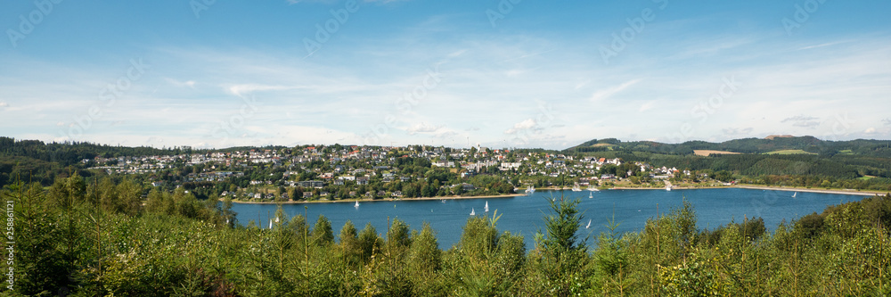 Wide view over Lake Sorpesee with sailboats and the dam with Langscheid in the background in front of a cloudy sky in Sundern, Sauerland, Germany