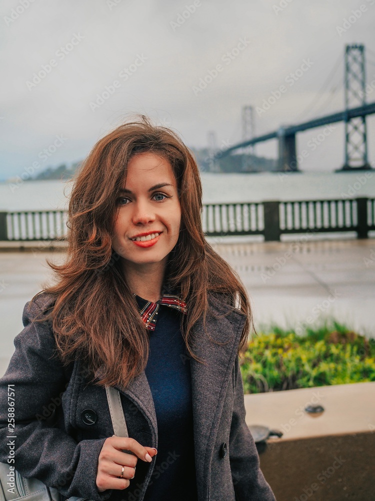 Brunette businesswoman girl in coat stands in front of Oakland Bay Bridge In San Francisco