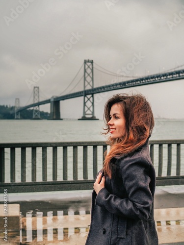 Brunette businesswoman girl in coat stands in front of Oakland Bay Bridge In San Francisco © KseniaJoyg