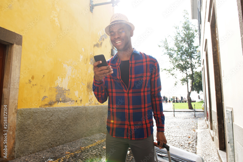 happy african american man looking at cellphone while walking in street with suitcase bag happy african american man looking at cellphone while walking in street with suitcase bag