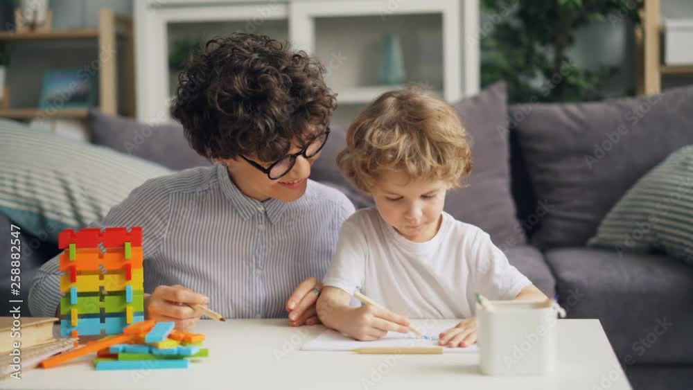 Creative little boy is drawing picture with pencils while cheerful woman is talking and smiling proud of her child. Family, creativity and leisure time concept. Creative little boy is drawing picture with pencils while cheerful woman is talking and smiling proud of her child. Family, creativity and leisure time concept.