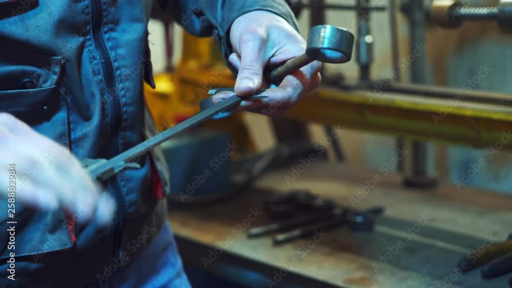 Mechanic is working with metal in a workshop, making measurements of metal parts. Worker holds a caliper in his hand, in a special suit on factory background. Mechanic is working with metal in a workshop, making measurements of metal parts. Worker holds a caliper in his hand, in a special suit on factory background.