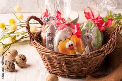 wicker brown basket with wrapped easter cookies near quail eggs and blossoming branch on wooden surface