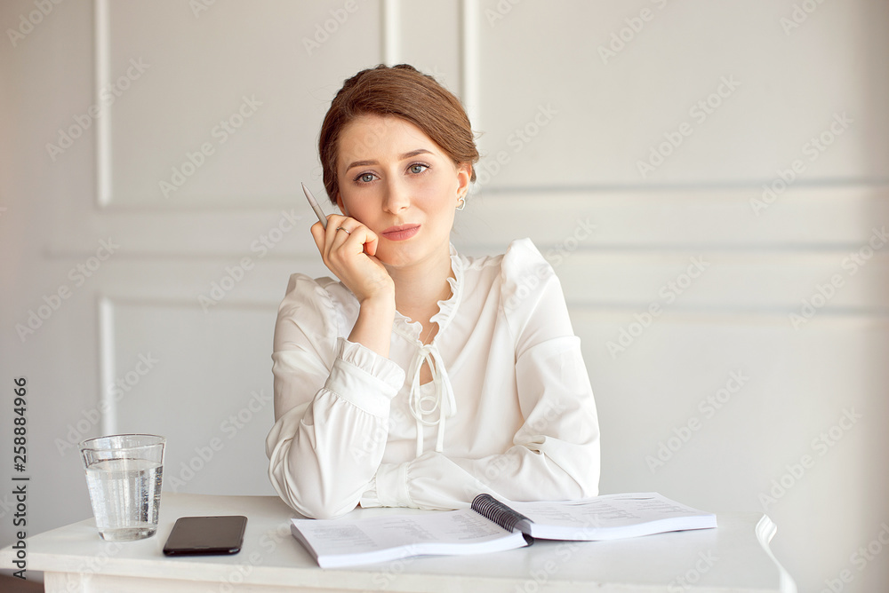 Portrait of a beautiful smiling young brunette businesswoman in a white shirt sitting on a bright modern workstation working with papers Portrait of a beautiful smiling young brunette businesswoman in a white shirt sitting on a bright modern workstation working with papers