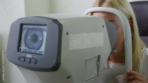 Tracking shot of cheerful blond woman looking into autorefractor machine during eye exam at optometrist photo
