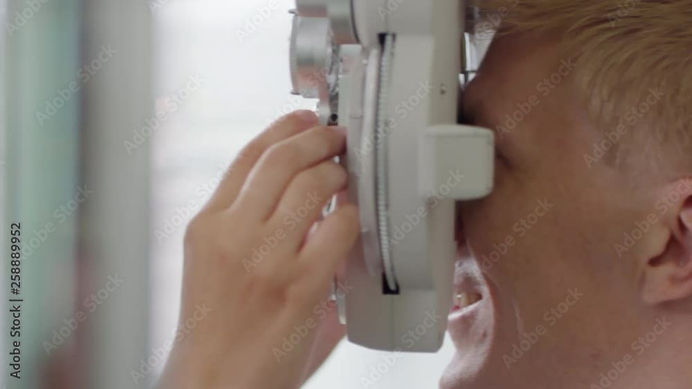 Tracking shot of cheerful man looking into phoropter and smiling as female optometrist changing lenses and asking questions Tracking shot of cheerful man looking into phoropter and smiling as female optometrist changing lenses and asking questions