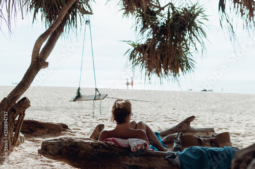 Back view of woman lying on the log and watching something in front of her on tropical beach with swing under the tree.