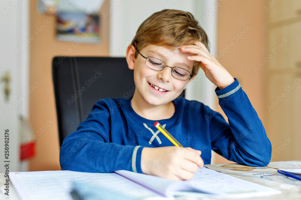 Fototapeta Portret śliczna szkolna dzieciak chłopiec jest ubranym szkła robi pracie domowej w domu. Mały skoncentrowany dziecka writing z kolorowymi ołówkami, indoors. Szkoła podstawowa i edukacja