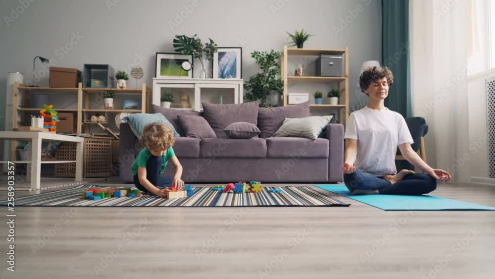 Small child is playing with blocks on floor while his young mommy meditating in lotus position on yoga mat. Relaxation, childhood and family house concept. Small child is playing with blocks on floor while his young mommy meditating in lotus position on yoga mat. Relaxation, childhood and family house concept.