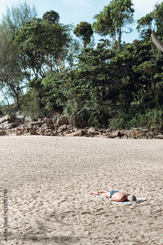 A lonely man lying on stomach sunbathing on the beach wearing a blue swimwear with a lot of tree in background.