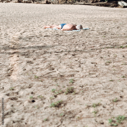 A lonely man lying on stomach sunbathing on the beach wearing a blue swimwear.