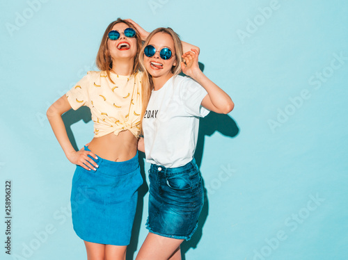 Two young beautiful smiling blond hipster girls in trendy summer jeans skirts clothes. Sexy carefree women posing near blue wall in round sunglasses. Positive models having fun and hugging