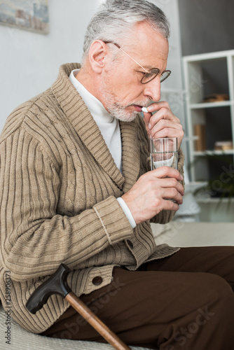 retired man taking pill while sitting on sofa near walking cane