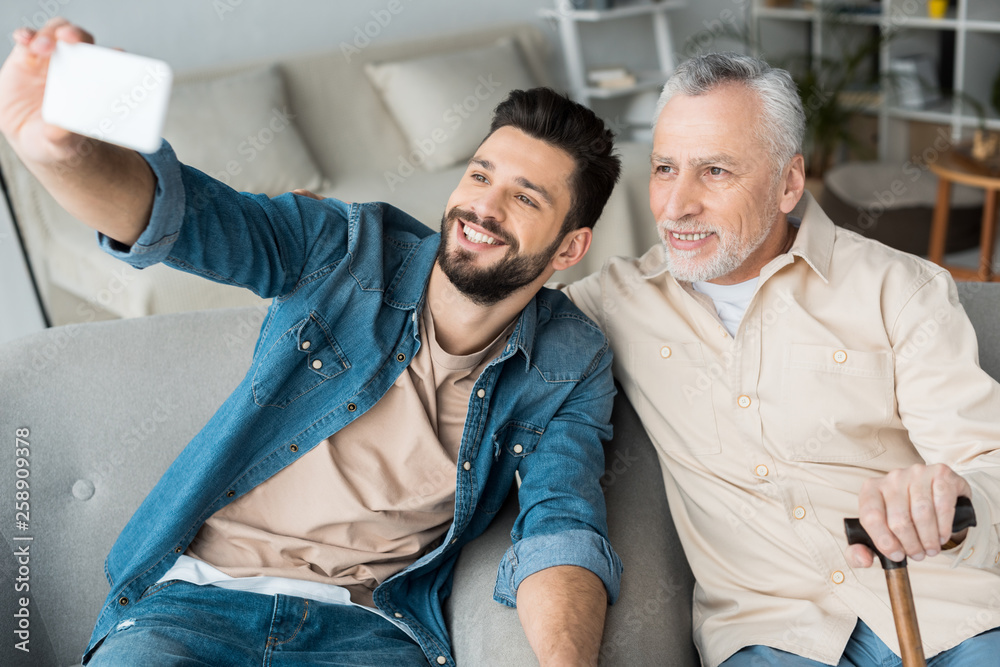 cheerful son taking selfie with retired cheerful father on smartphone cheerful son taking selfie with retired cheerful father on smartphone