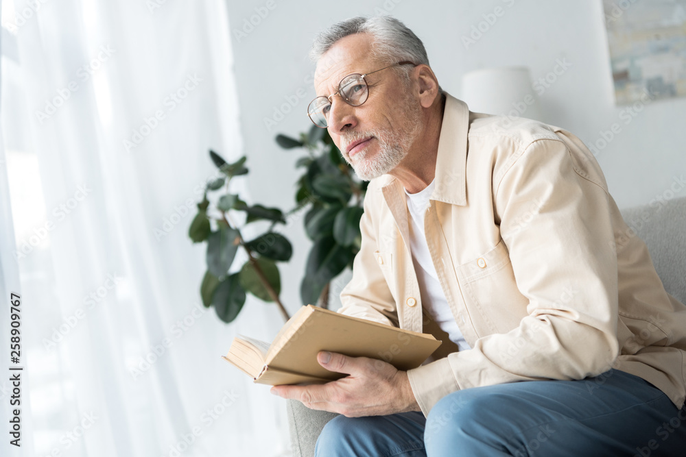 senior man in glasses holding book while sitting at home