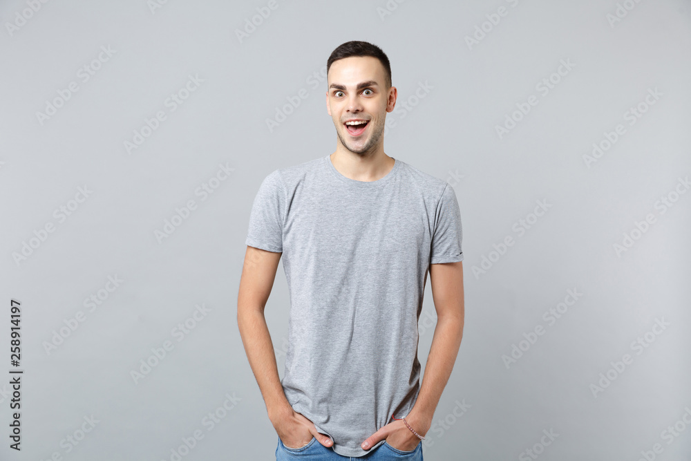 Portrait of excited young man in casual clothes keeping mouth open, holding hands in pockets isolated on grey wall background in studio. People sincere emotions, lifestyle concept. Mock up copy space. Portrait of excited young man in casual clothes keeping mouth open, holding hands in pockets isolated on grey wall background in studio. People sincere emotions, lifestyle concept. Mock up copy space.