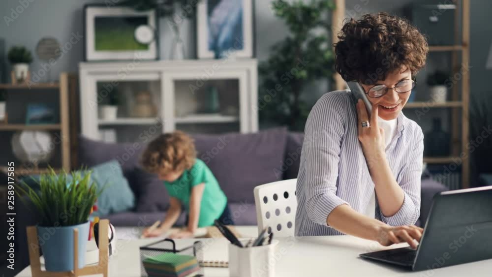 Busy girl is talking on mobile phone and working with laptop typing while her small son is playing at home. Business people, family and house concept.