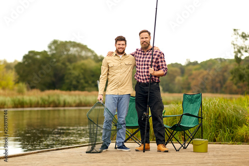 leisure and people concept - happy male friends with fishing rod and scoop net hugging at lake or river