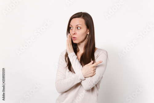 Attractive young woman whispers gossip and tells secret with hand gesture, pointing index finger aside isolated on white wall background. People sincere emotions lifestyle concept. Mock up copy space.