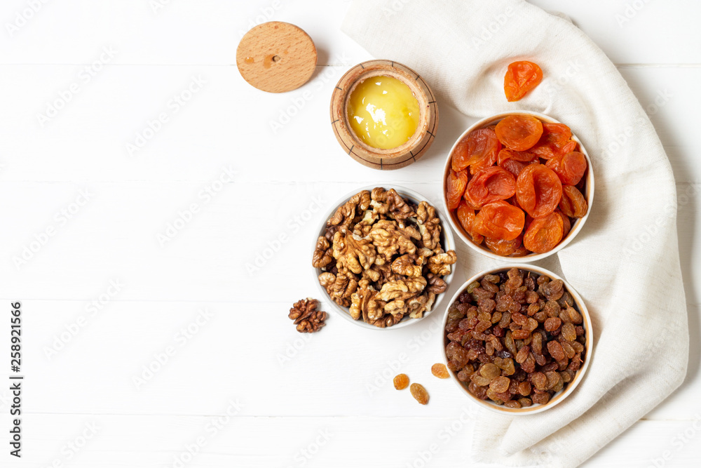 Obraz premium Dried apricots, raisins, walnuts in cups and a barrel of honey standing on a napkin on a white wooden background.
