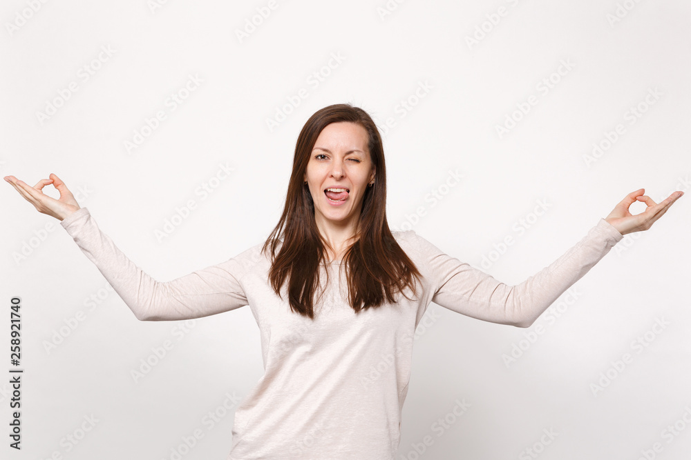 Portrait of funny blinking young woman in light clothes hold hands in yoga gesture showing tongue isolated on white background in studio. People sincere emotions lifestyle concept. Mock up copy space. Portrait of funny blinking young woman in light clothes hold hands in yoga gesture showing tongue isolated on white background in studio. People sincere emotions lifestyle concept. Mock up copy space.